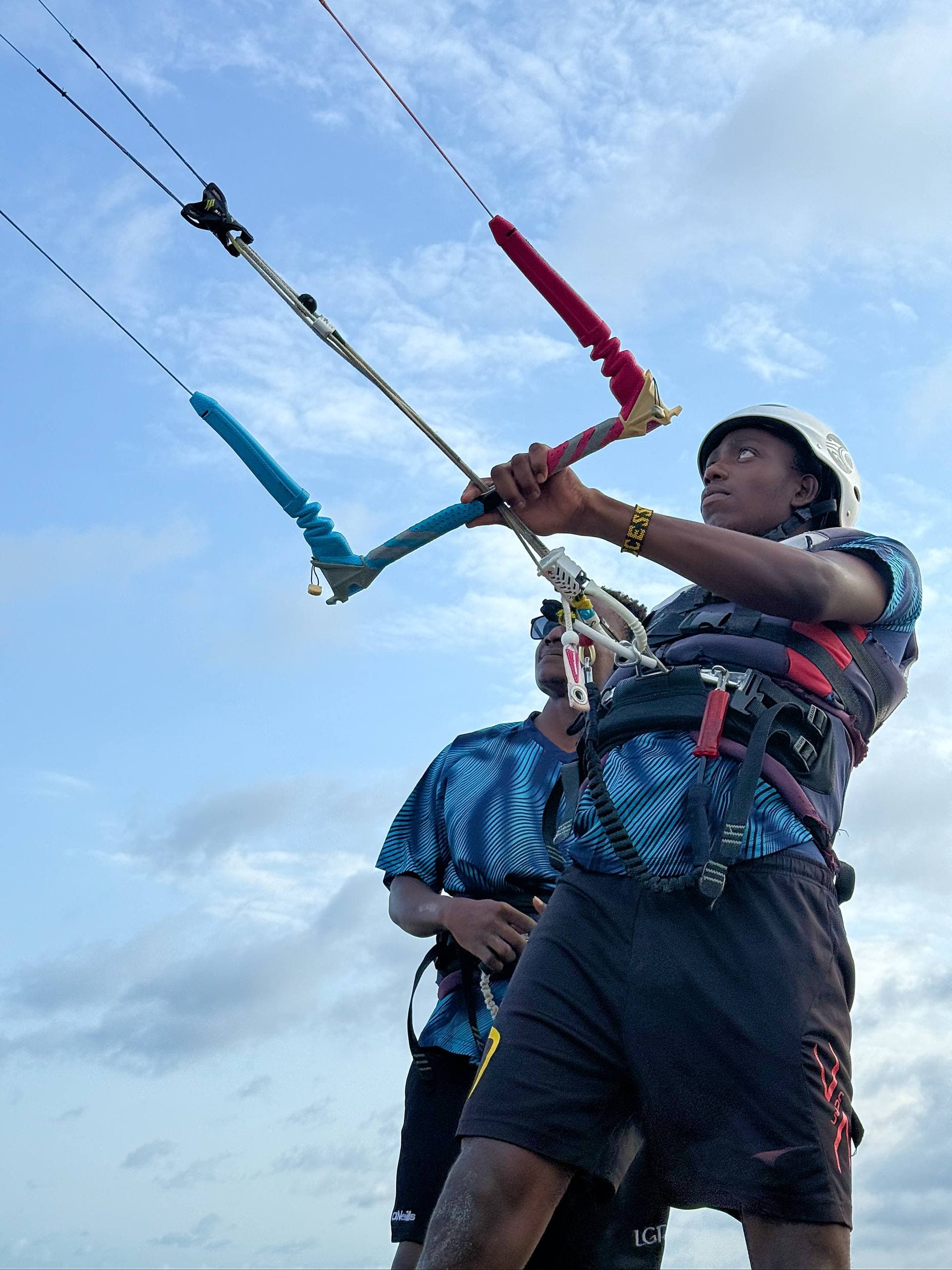 A young rider in a helmet and harness grips the kite control bar and looks up at the kite, an instructor guiding from close behind, against a blue Watamu sky